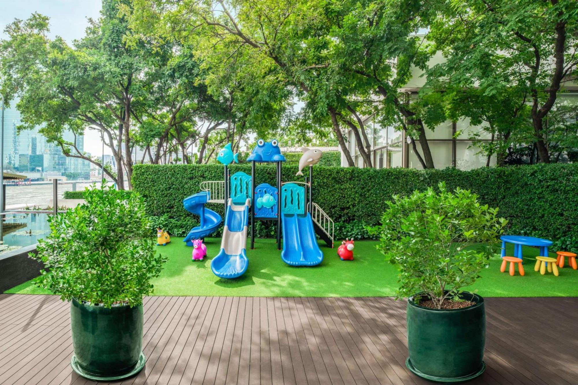 Millennium Hilton Bangkok Hotell Eksteriør bilde The photo features a playground area surrounded by greenery. In the center, there are blue slides and climbing structures designed for children, resembling friendly animals. The playground is set on a grassy surface, and there are colorful toys and f