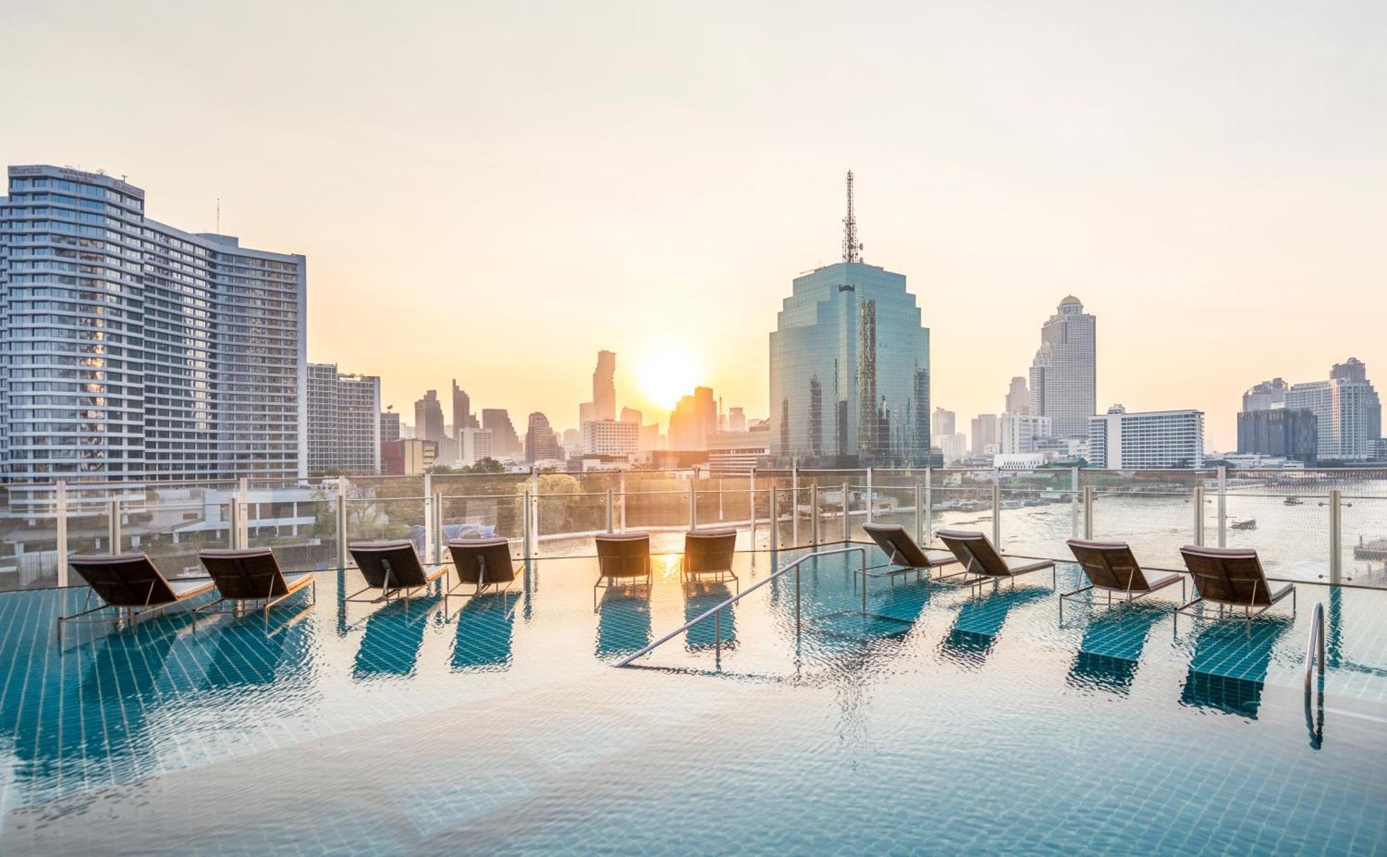 Millennium Hilton Bangkok Hotell Eksteriør bilde The photo depicts a beautiful rooftop swimming pool with lounge chairs positioned around its edge. The pool reflects the colors of a vibrant sunset, casting a warm glow over the scene. In the background, a skyline of modern buildings can be seen, inc
